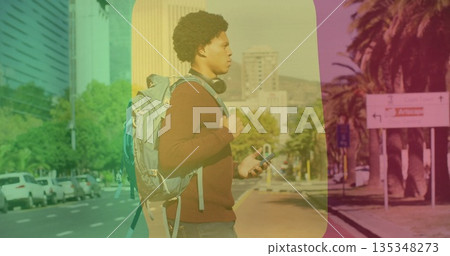 Standing man wearing maroon sweater, jeans on sidewalk, holding smartphone, backpack and headphones 135348273