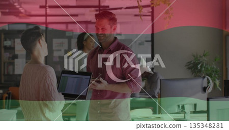 Presenting woman showing data on laptop to coworker with smartphone in open-plan office with plants 135348281