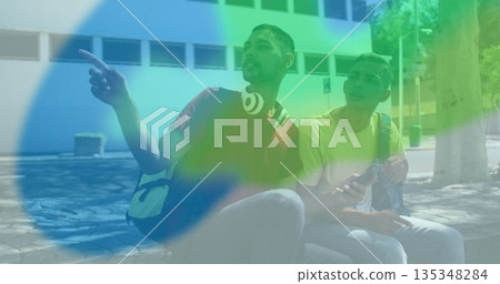 Pointing adult men sitting and looking left on bench at courtyard, with backpacks and headphones Pointing adult men sitting and looking left on bench at courtyard, with backpacks and headphones 135348284