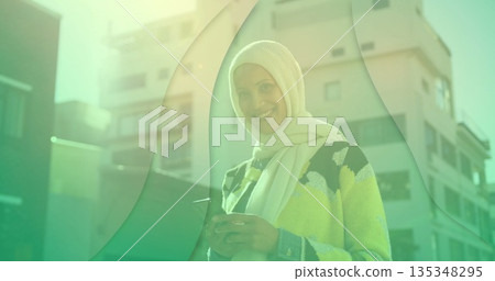 Mature Middle Eastern woman wearing hijab holding smartphone in outdoor plaza with green overlay Mature Middle Eastern woman wearing hijab holding smartphone in outdoor plaza with green overlay 135348295