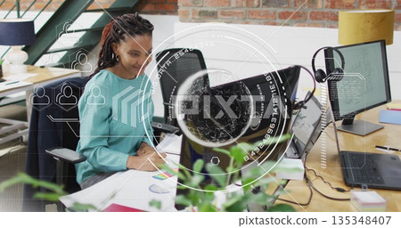 Woman in teal blouse sketching chart and viewing code on laptop in open-plan office, with monitors 135348407