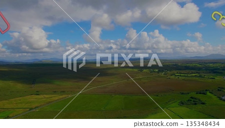 Narrow country road snaking through green fields in aerial view, with hedgerows, hills, clouds 135348434