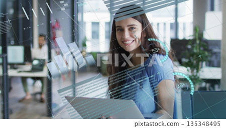 Working woman sitting at desk smiling in open-plan office, with laptop, holographic data overlays Working woman sitting at desk smiling in open-plan office, with laptop, holographic data overlays 135348495
