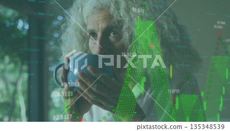 Sipping mature woman holding blue mug and reviewing candlestick charts at window, data overlays 135348539
