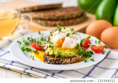 Toast with avocado and egg with cup of tea for healthy breakfast or snack. Sandwich with avocado and poached egg on a white table, selective focus. Toast with avocado and egg with cup of tea for healthy breakfast or snack. Sandwich with avocado and poached egg on a white table, selective focus. 135349312