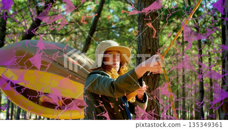 Woman wearing bucket hat and jacket tightening rope in forest, with hammock, purple digital overlay 135349361