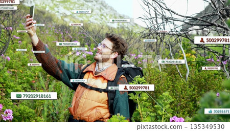 Holding smartphone, hiker in orange vest taking selfie on rocky slope with backpack, digital labels 135349530