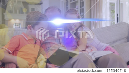 Reading grandfather sharing open book with grandchildren on sofa in living room, with fireplace Reading grandfather sharing open book with grandchildren on sofa in living room, with fireplace 135349748