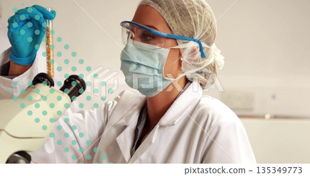 Examining scientist holding sample tube in gloved hand at eye level on lab bench, with microscope Examining scientist holding sample tube in gloved hand at eye level on lab bench, with microscope 135349773