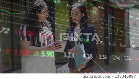 Comparing stock charts on tablet and folder by businesswomen in office, displaying market numbers Comparing stock charts on tablet and folder by businesswomen in office, displaying market numbers 135349887
