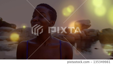 Woman wearing swimsuit standing in calm water at rocky shore, with boulders and lens flares 135349981