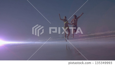 Running and leaping couple wearing swimsuit and trunks on wet sand with lens flare, ocean waves Running and leaping couple wearing swimsuit and trunks on wet sand with lens flare, ocean waves 135349993
