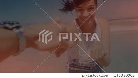 Smiling woman wearing white print top and denim shorts reaching forward at sandy beach, with watch 135350002