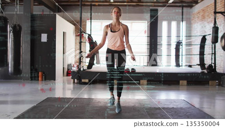 Jumping woman skipping rope on foam mats in boxing studio, with punching bags and boxing ring 135350004