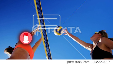 Jumping female volleyball players wearing sports tops hitting volleyball over net on beach court Jumping female volleyball players wearing sports tops hitting volleyball over net on beach court 135350006