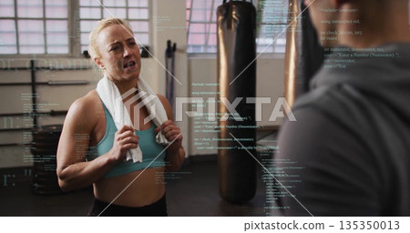 Woman wearing sportswear catching breath in fitness studio, with punching bag and code overlays 135350013