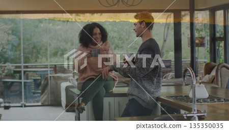 Leaning man and sitting woman holding mugs on island in modern kitchen, showing stovetop and faucet 135350035