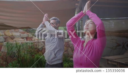 Practicing couple in sportswear performing yoga prayer pose on garden deck, with canopy and pond 135350036