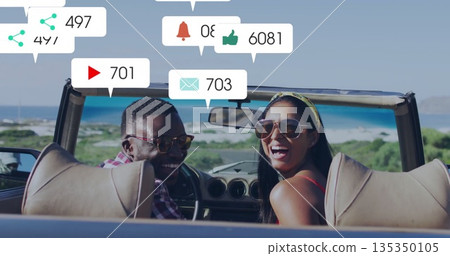 Sitting couple wearing sunglasses, viewing floating social icons in convertible car on coastal road 135350105