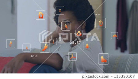 Sitting mid adult woman leaning on sofa arm in living room, with floating energy usage icons 135350107