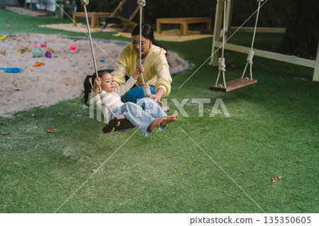 cheerful child girl with mother playing on wooden swing at playground cheerful child girl with mother playing on wooden swing at playground 135350605