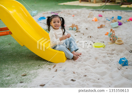 child girl playing and sitting at the bottom of yellow slide in a sandy playground. 135350613