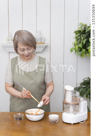 一位老年婦女在廚房裡用食物料理機做飯。 135350694
