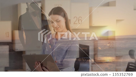 Businesswoman wearing blouse using laptop in office with sunset ocean panels and man in fedora coat Businesswoman wearing blouse using laptop in office with sunset ocean panels and man in fedora coat 135352769