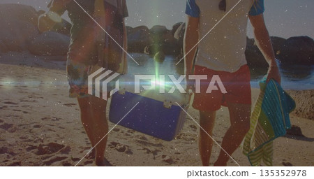 Walking two male friends carrying portable cooler and striped towel on sandy shoreline at sunset Walking two male friends carrying portable cooler and striped towel on sandy shoreline at sunset 135352978
