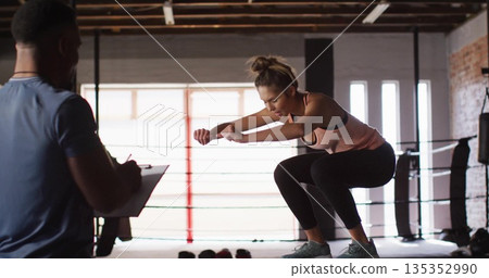Jumping woman in sport gear performing squat jump in boxing ring with trainer clipboard, copy space Jumping woman in sport gear performing squat jump in boxing ring with trainer clipboard, copy space 135352990