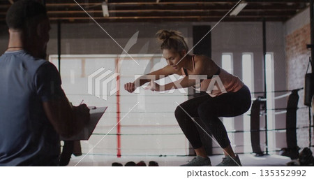 Trainee in sportswear performing squat jump onto step in boxing ring, trainer holding clipboard Trainee in sportswear performing squat jump onto step in boxing ring, trainer holding clipboard 135352992