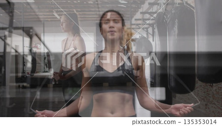 Jumping brown woman holding jump rope in fitness studio, with punching bags, mirrors and dumbbells Jumping brown woman holding jump rope in fitness studio, with punching bags, mirrors and dumbbells 135353014