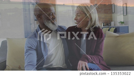 Smiling senior couple sitting closely on sofa in living room, with large window sunset view 135353022