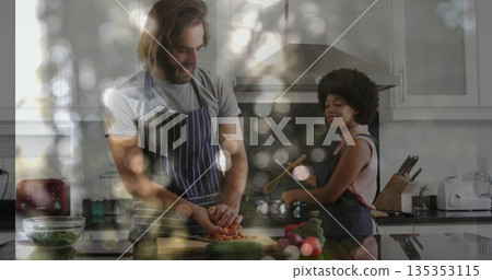 Man wearing apron slicing carrots and woman stirring pot with wooden spoon in kitchen with greens Man wearing apron slicing carrots and woman stirring pot with wooden spoon in kitchen with greens 135353115