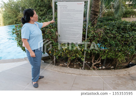 Woman points at a sign near a pool area in a sunny location while wearing casual clothing 135353595