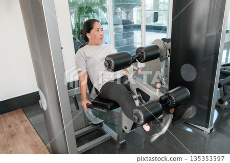 Woman exercises in gym using leg press machine during morning hours in bright workout room with large windows Woman exercises in gym using leg press machine during morning hours in bright workout room with large windows 135353597