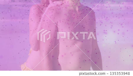 Embracing pair wearing dark swim trunks and yellow bikini bottom at beach shoreline, pink overlay 135353948