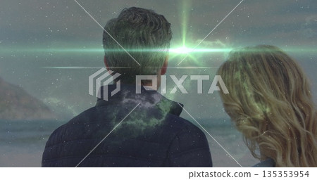 Gazing mature couple wearing jackets standing on sandy shore, with cosmic double exposure effect 135353954