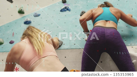 Two women wearing sports bras and leggings climbing on climbing wall at gym, with colorful holds 135353965