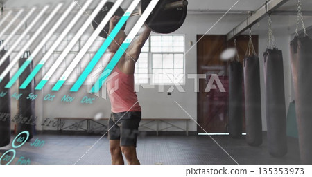 Pressing man in red tank top lifting barbell overhead in gym with punching bags, chart overlay Pressing man in red tank top lifting barbell overhead in gym with punching bags, chart overlay 135353973