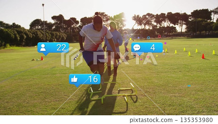 Jumping African American female player wearing jersey clearing hurdle on grass field, with cones 135353980