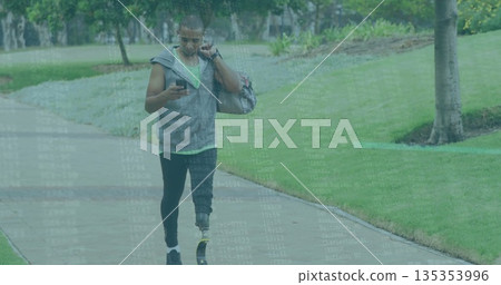 Walking athlete in hoodie talking on smartphone on park path with duffel bag and prosthetic blade Walking athlete in hoodie talking on smartphone on park path with duffel bag and prosthetic blade 135353996
