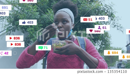 Eating woman wearing pink sweater and blue mask on sidewalk, holding salad with social media icons 135354110