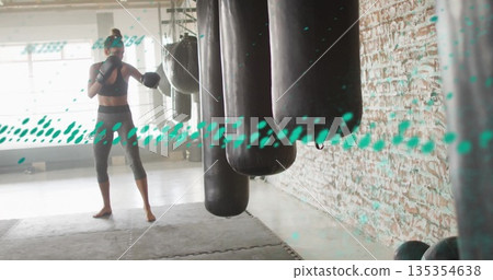 Preparing boxer in sports bra holding fighting stance in boxing gym, with punching bags and gloves 135354638