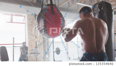 Punching shirtless boxer wearing boxing gloves hitting bag in brick gym, with squat rack Punching shirtless boxer wearing boxing gloves hitting bag in brick gym, with squat rack 135354640