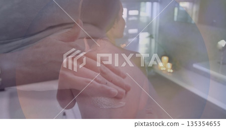 Massaging therapist applying cream to client's shoulders on spa bed, with candles, towel and lotion 135354655