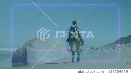 Walking man with prosthetic leg along sandy shoreline, with backpack, board and camera tripod 135354659