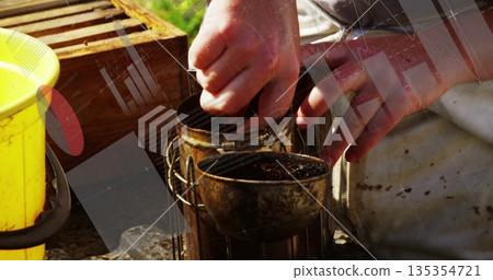 Operating beekeeper handling metal bee smokers at apiary, wooden hive box frames and yellow bucket Operating beekeeper handling metal bee smokers at apiary, wooden hive box frames and yellow bucket 135354721