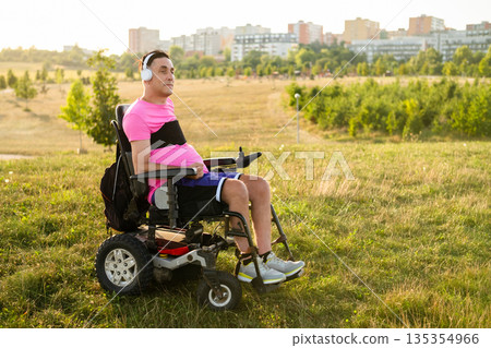 Relaxed male sitting in a wheelchair enjoying favorite music with white headphones outdoors in sunlight. 135354966