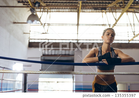 Female athlete leaning on boxing ropes under natural light in gym with hand wraps, copy space 135355397